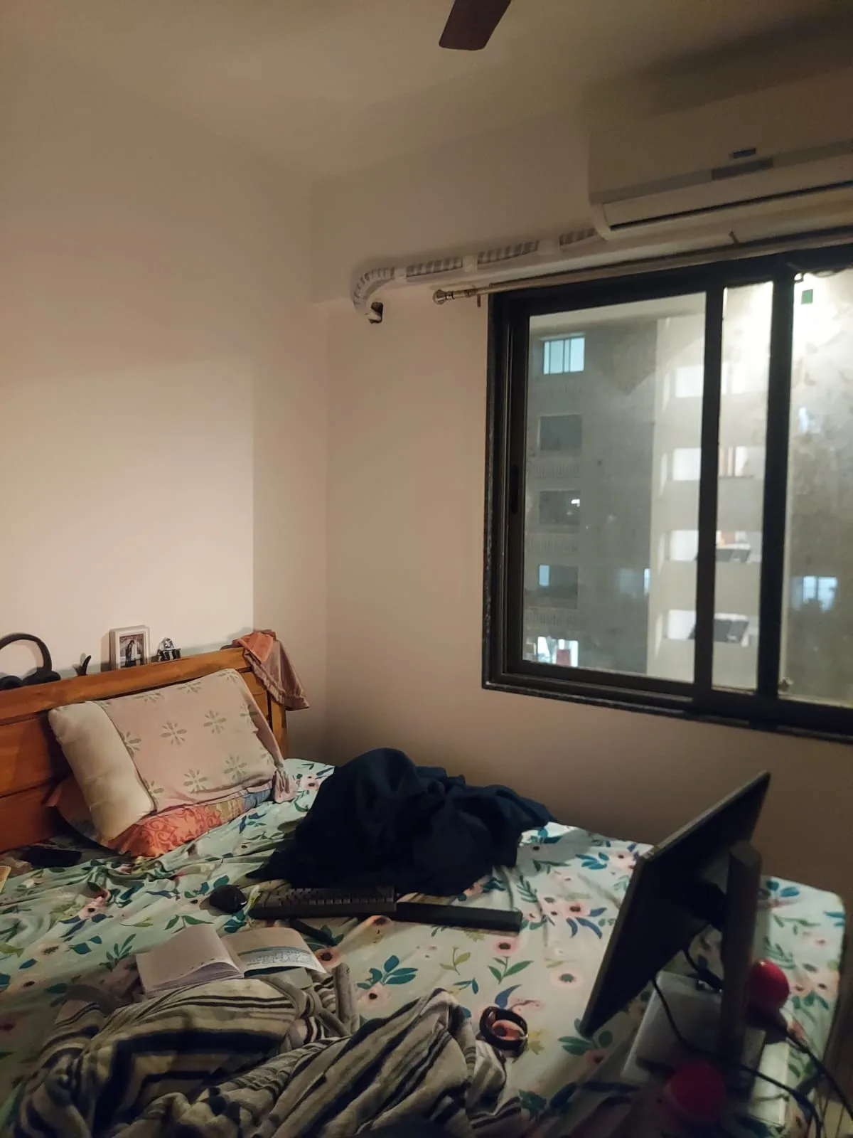 A monitor, keyboard and mouse set up directly on a floral-sheeted bed next to a window at night.
