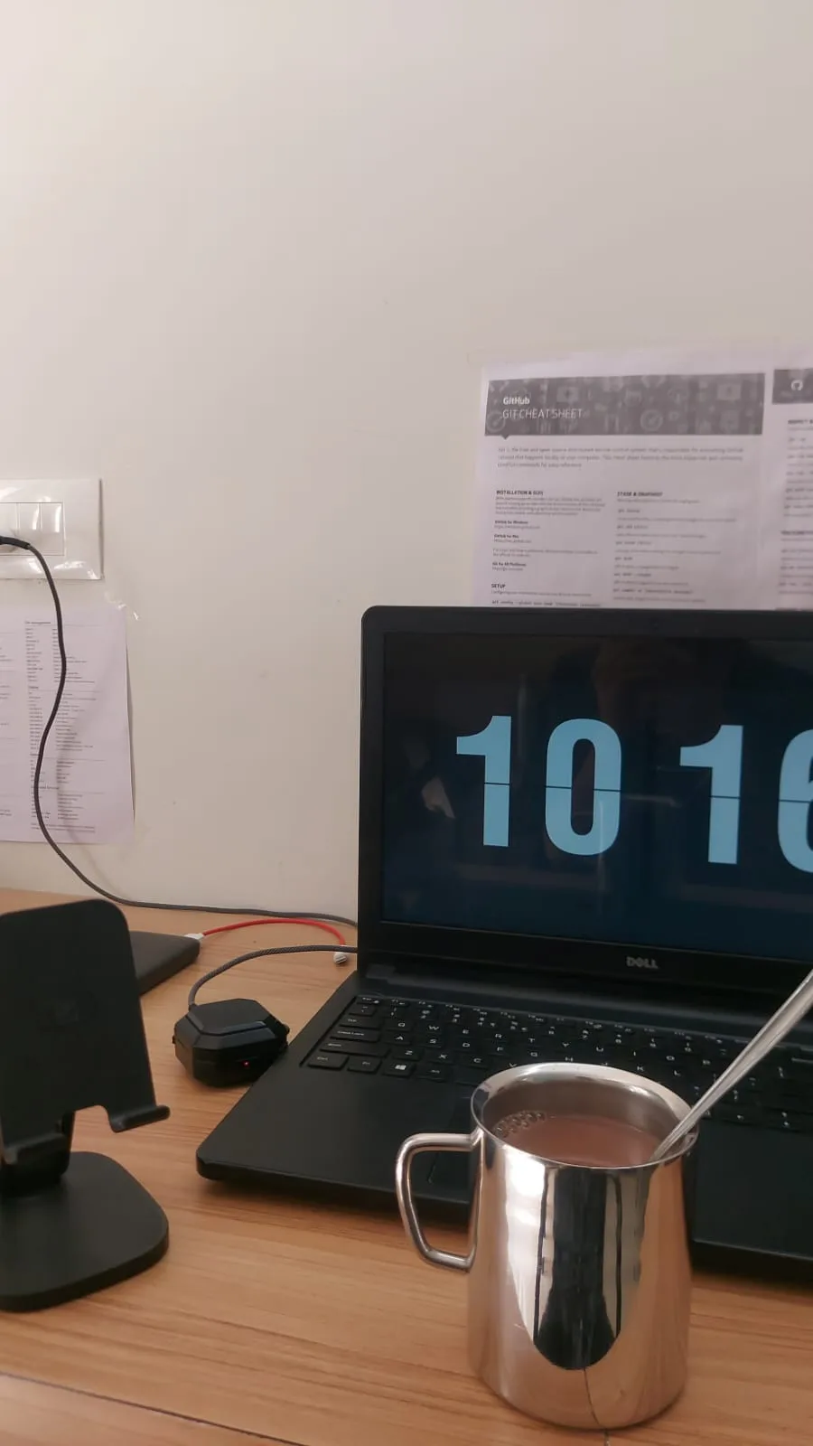 The Dell on a desk with a flip clock, a git cheat sheet taped to the wall, a phone stand and a mug.