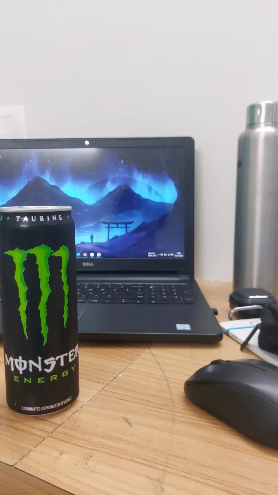 The Dell laptop with an aurora wallpaper, next to a Monster energy can and a flask.