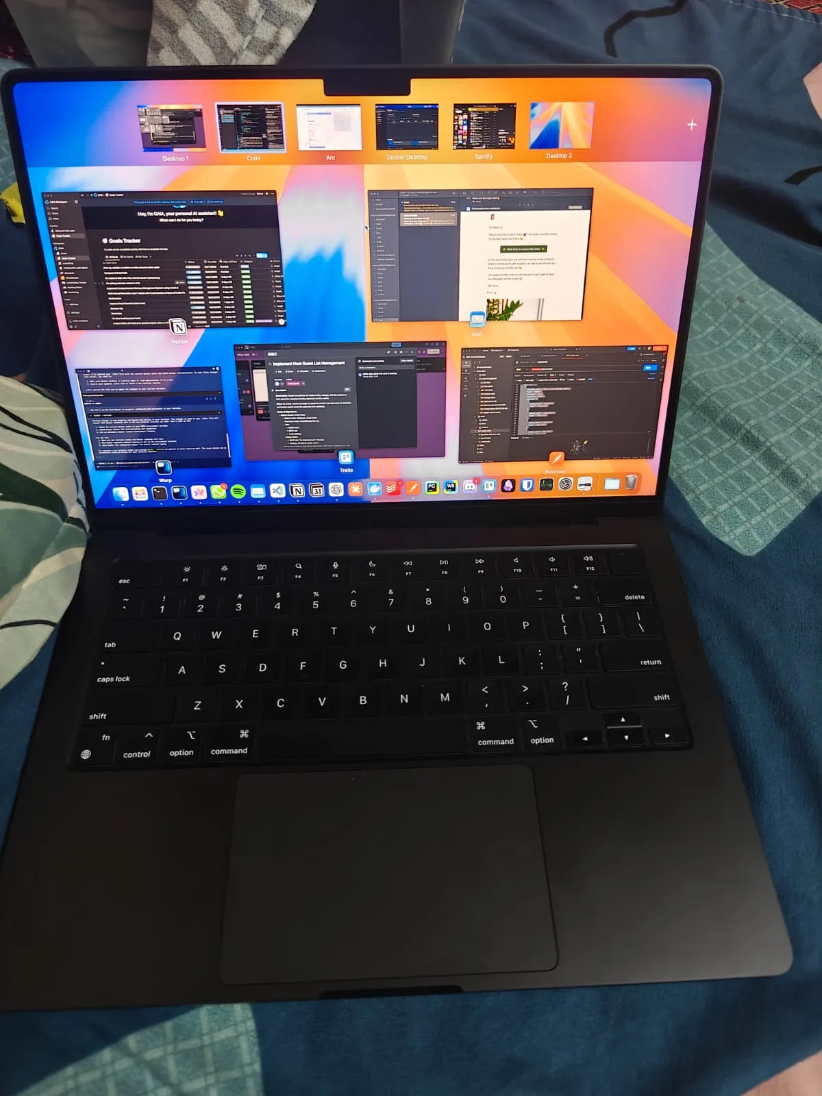 A brand new black MacBook on a bed showing the macOS Mission Control view with GAIA, Notion, Mail, Warp, Trello and Postman windows open.