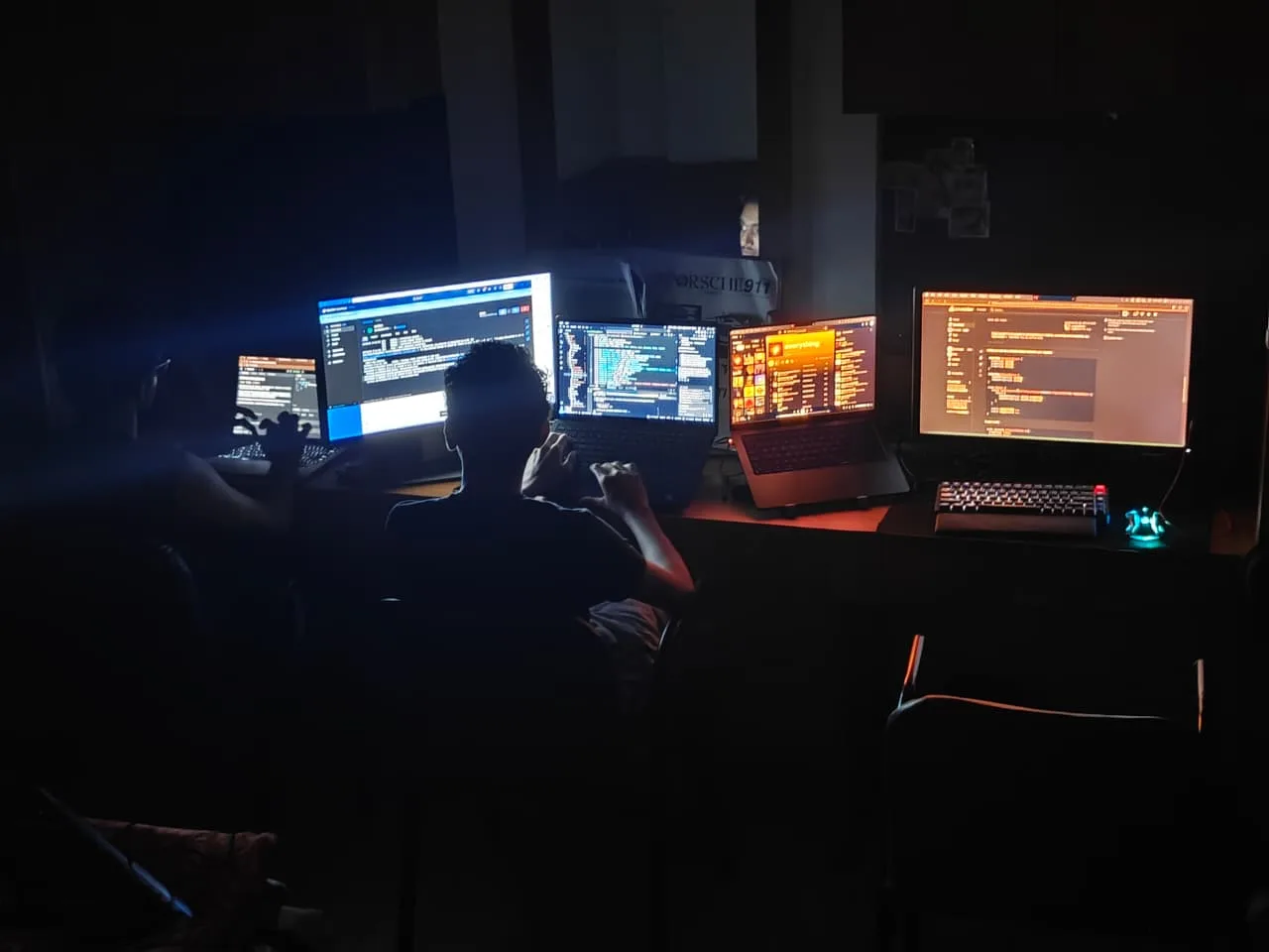 A dark room with a multi-monitor desk all lit up with code and a Porsche 911 chair cover, working late into the night.
