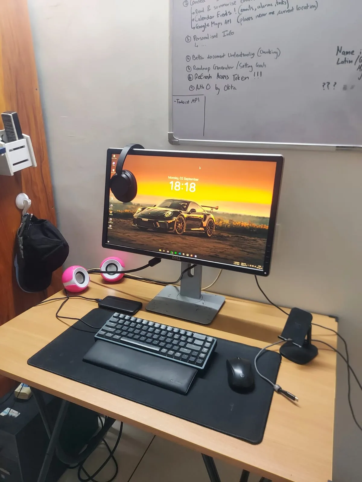 The Dell monitor on the new table with a Porsche 911 wallpaper, a Keychron keyboard, headphones, and a whiteboard full of GAIA roadmap notes behind it.