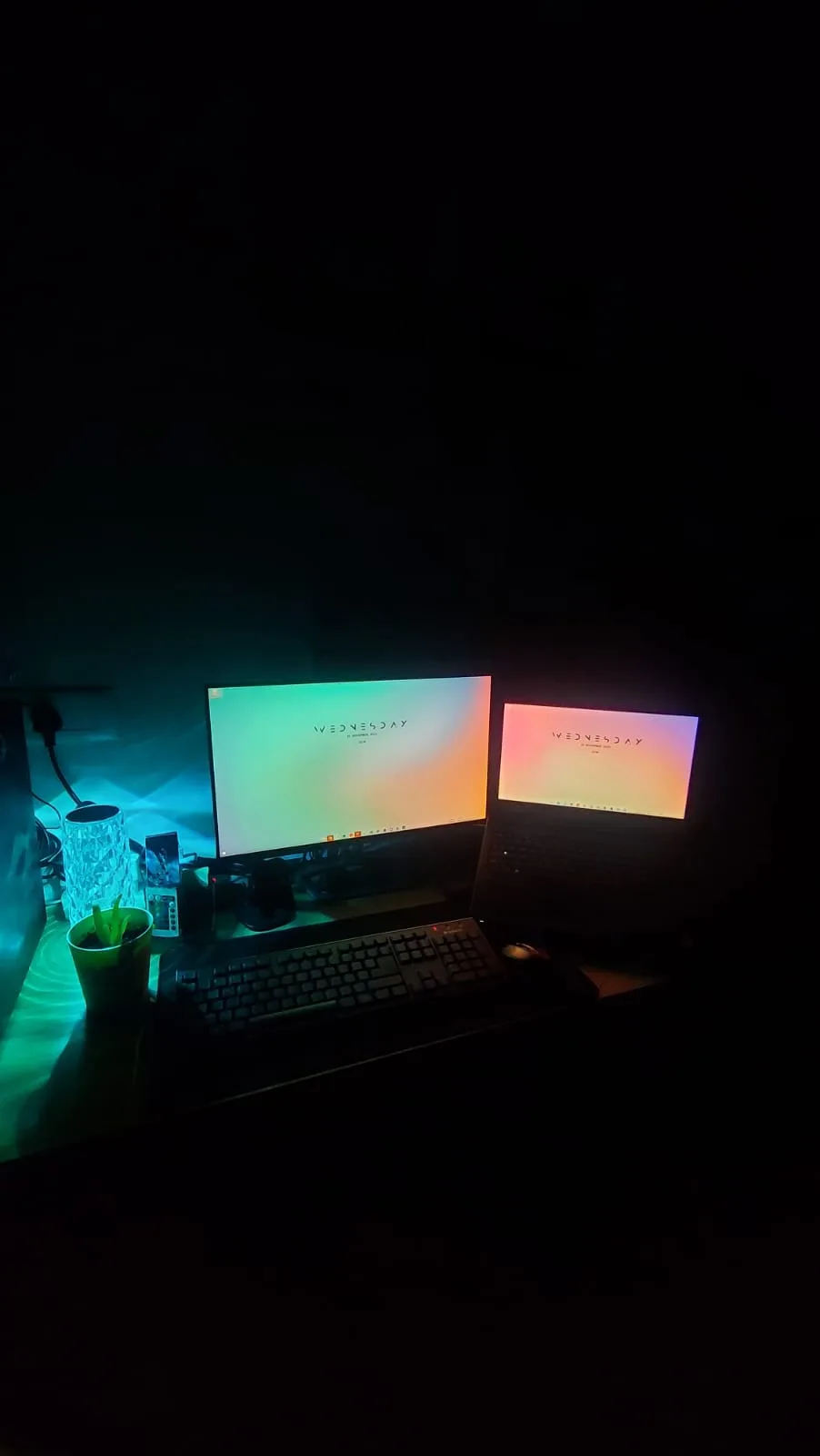 The monitor and Dell laptop at night with an RGB lamp glowing teal and a 'Wednesday' wallpaper.