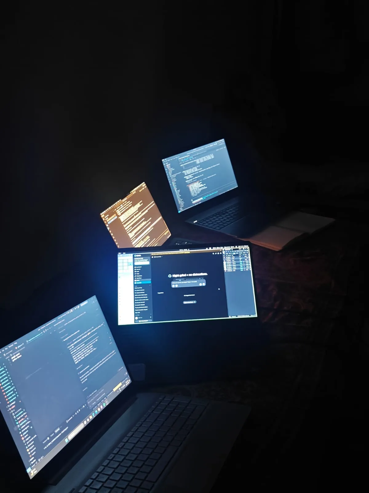 Four laptops glowing with code in a dark room, all lined up on a bed, during the depressing tiny-room job phase.