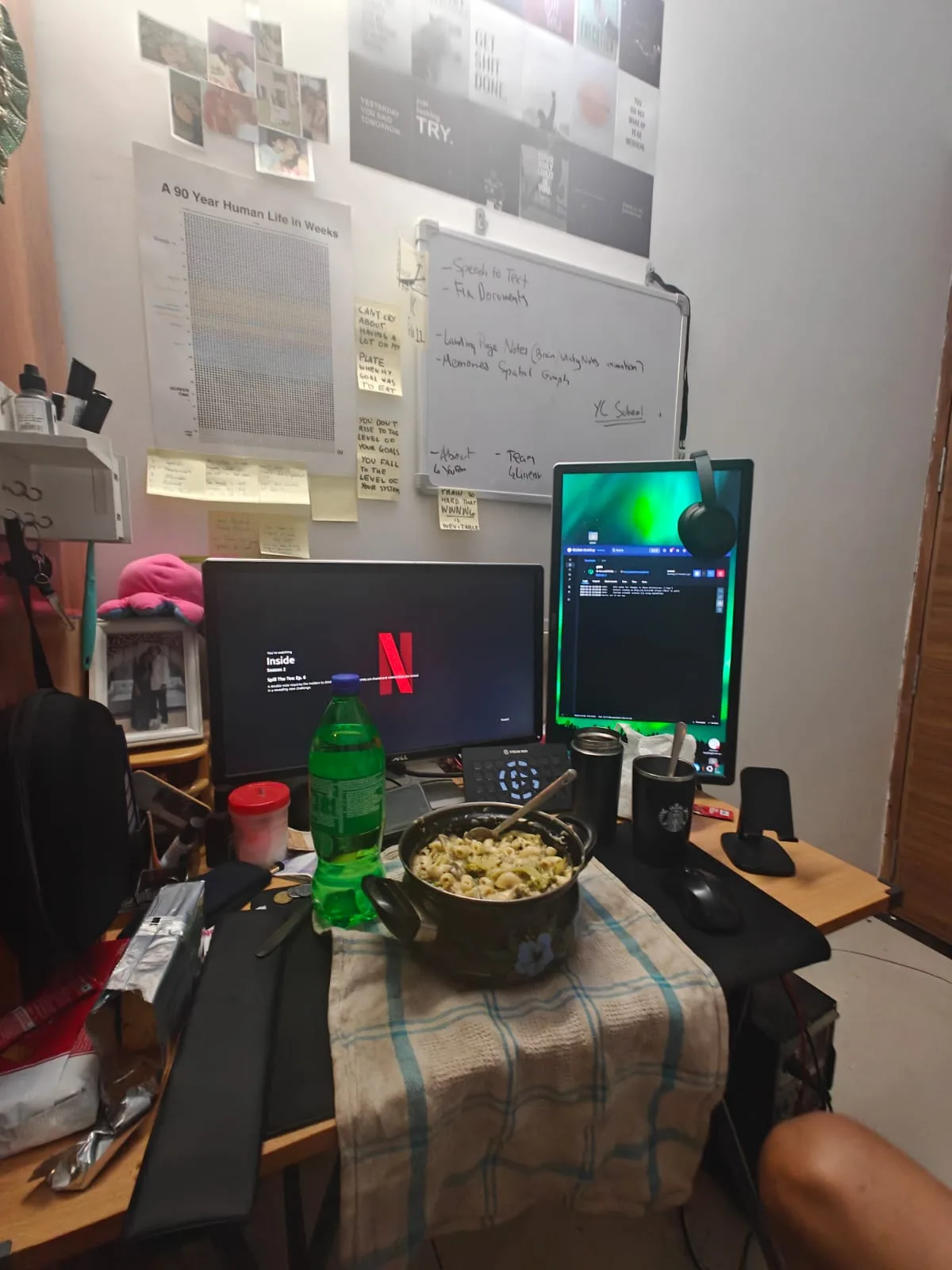 A busy desk with a vertical second monitor, Netflix playing on the main screen, a bowl of pasta, and a wall full of motivational posters and whiteboard notes.